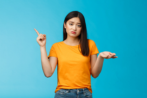 Troubled cute upset asian girl have problem asking your help, pointing upper left corner shrugging unsure, look frustrated and sad, questioned answer, cannot deal problem, stand blue background.