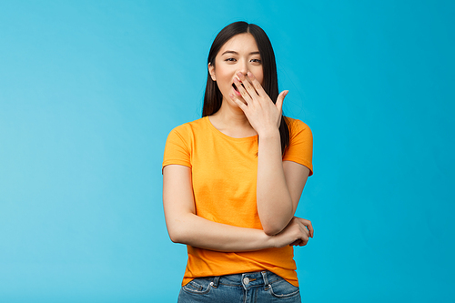 Sleepy cute asian brunette yawning waiting for cup coffee morning standing office smiling upbeat, cover opened mouth waking up feel energized, stand blue background. Copy space