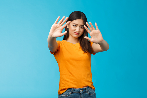 Asian moody displeased silly timid girl asking turn off light not photograph her, raise hands block, cover face step back and cringe disappointed, standing blue background upset reluctant. Copy space