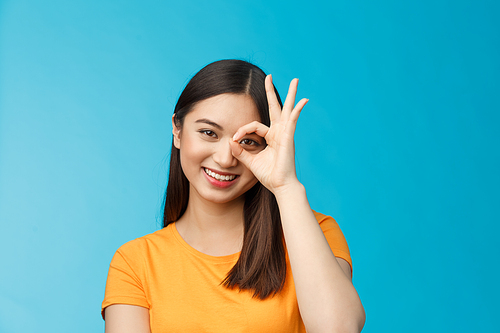 Close-up cute dreamy tender asian woman dark short haircut show okay ok sign look through circle smiling broadly, express positivity good mood, standing upbeat friendly gaze camera, blue background.