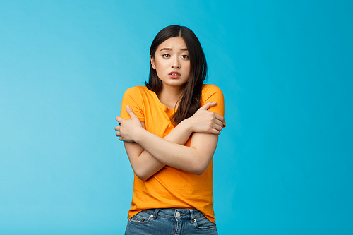 Girl feeling uncomfortable walking light yellow t-shirt, hugging herself trembling, shaking feeling cold, freezing windy weather, frowning grimacing discomfort, stand blue background.