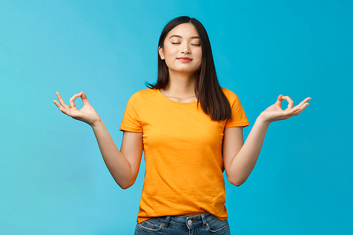 Peaceful charming relaxed asian girl buddhist meditating, breathing, inhale fresh air practice yoga, close eyes smiling relieved, stand lotus nirvana pose, reach zen, standing blue background.