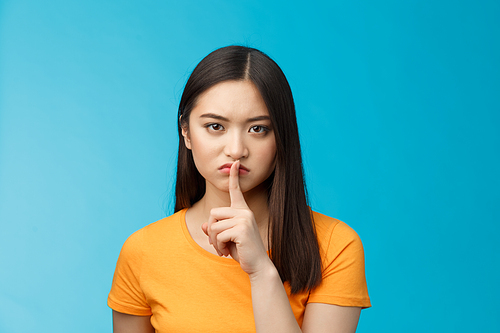 Hush stay silent. Serious-looking asian girl look displeased grumpy show shush sign press index finger lips, frowning upset, telling not speak, untell secret, prohibit loud music, blue background.