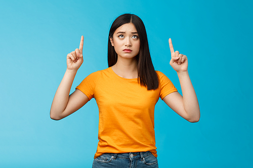 Upset gloomy cute asian young girl dark haircut frowning pointing looking up uneasy, feel regret cannot effort buy cool thing, stand blue background unsatisfied frustrated, wear yellow t-shirt.