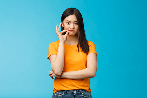 Serious-looking asian female friend promise keep secret, seal lips, hold zip near mouth look focused determined, stay silent and speechless, stand wearing yellow t-shirt blue background.