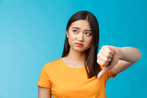 Disappointed female asian visitor give negative feedback, look away disturbed and upset, show thumb down dislike sign, judging bad thing, poor quality, stand blue background. Copy space