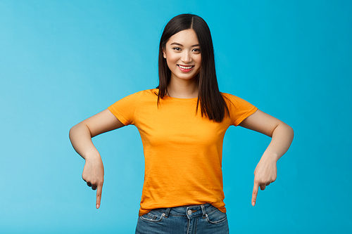 Cheerful self-assured asian girl dark haitcut wear yellow summer t-shirt introduce promo, pointing hands down, indicate bottom advertisement smiling delighted, share good link blue background.