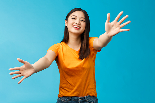 Friendly clingy girlfriend walking towards camera with extended hands to give tight hug, smiling broadly embracing friend, happily cuddling welcoming girlfriend, stand blue background.