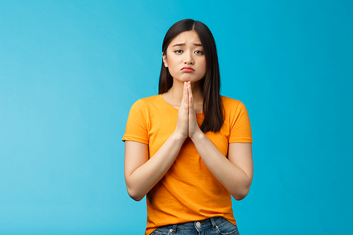 Upset cute silly asian girl praying, plead for help, pouting frowning need, make pitty face, hold hands pray begging for favor, apologizing feelings guilty sad, stand blue background.