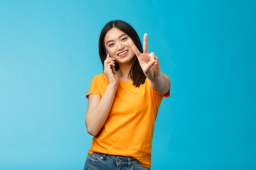 Charismatic cute girl share positive news talking smartphone show peace, victory sign smiling satisfied, standing blue background receive pleasant phone call, achieve deal via phone-call.