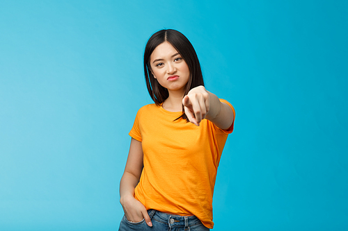 Disappointed attractive asian girl frowning, grimasing upset, pointing index finger camera upset unsatisfied, disliking you, accuse someone, blame person, standing displeased blue background.