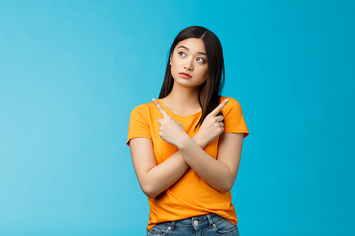 Confused cute asian female student thinking what do, look sideways thoughtful, cross hands chest pointing left and right, making choice, thinking what pick, taking important decision blue background.