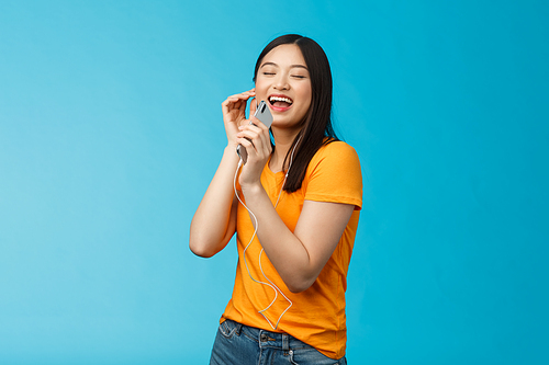 Joyful carefree attractive asian woman close eyes pleasure and joy, smiling wear earphones, listening music via headphones, hold smartphone like microphone karaoke, singing along favorite song.