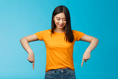 Cute tender feminine asian girl dark haircut look pointing down contemplating interesting product, stare curiously smiling amused, express enthusiasm interest try-out cool thing, blue background.