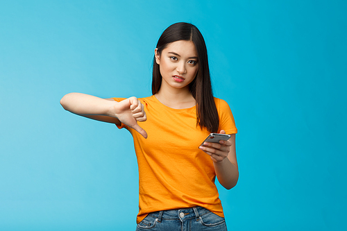 Displeased asian woman short dark haircut, frowning grimacing disappointed, show thumb down disapprove bad app, slow wifi speed hold smartphone unsatisfied, stand blue background. Technology concept