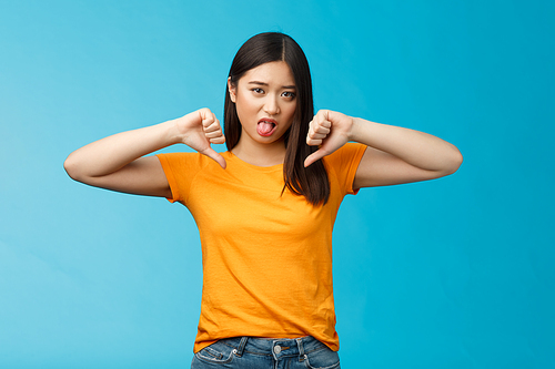 Disappointed upset and unimpressed cute childish asian girl dark haircut, frowning cringing dislike, aversion, show thumbs-down stick tongue disgust, give negative opinion judging, blue background.