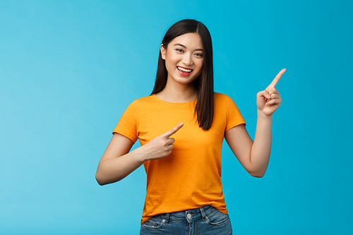 Cheerful lively cute asian woman smiling laughing upbeat, pointing upper right corner, give advice what choose, shopping online, grinning happily, enthusiastic promo, blue background.