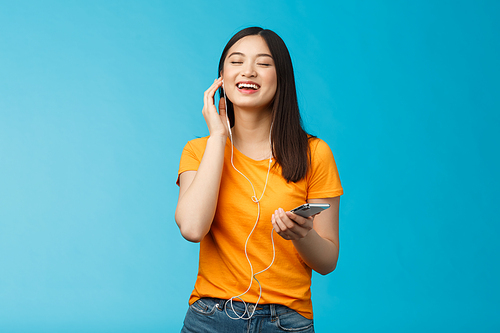 Lovely carefree cute asian girl enjoy awesome music boost mood listening favorite songs, touch earphone close eyes from pleasure delight, singing along, hold smartphone stand blue background.