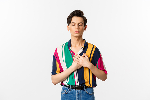 Image of nostalgic young gay man standing with closed eyes and hands on heart, remember something, standing over white background.