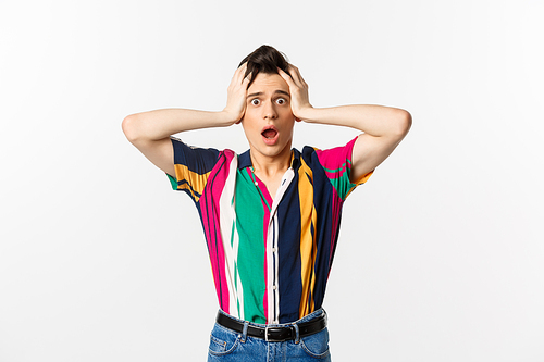 Scared and worried young gay man panicking, holding hands on head and looking alarmed at camera, standing over white background.