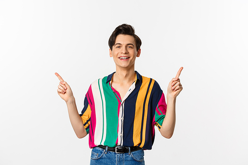 Happy young gay man pointing fingers sideways, showing two choices and smiling, standing against white background. Copy space