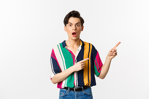 Impressed young man showing advertisement, open mouth with disbelief and pointing at upper right corner, standing against white background.