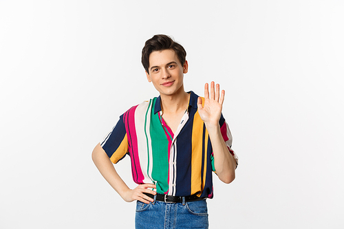Friendly young man smiling and saying hello, waving hand in greeting gesture, standing over white background.