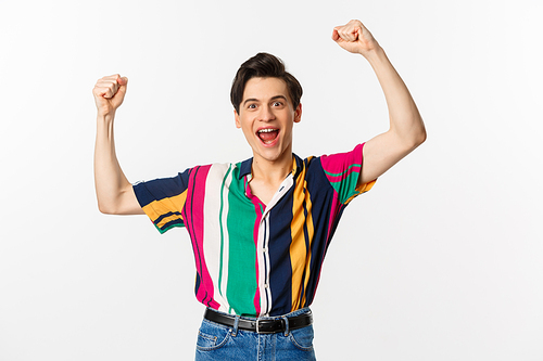 Image of happy young man triumphing of winning, celebrating victory, raising hands up in rejoice and shouting yes, standing over white background.