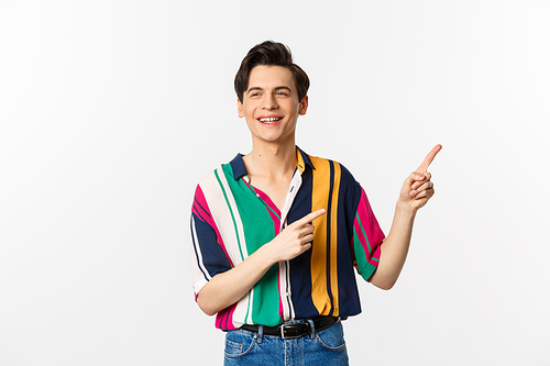 Carefree guy in stylish shirt pointing fingers at upper right corner, showing promo banner or logo, smiling and talking to someone, standing over white background.