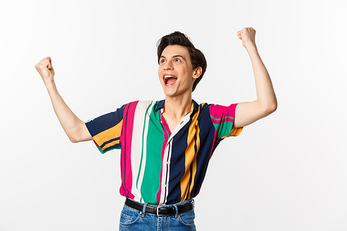 Happy gay man celebrating victory, raising hands up and rejoicing of winning, tirumphing while standing over white background.