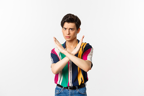 Disappointed young queer man telling no, showing stop gesture and forbid something bad, standing over white background.