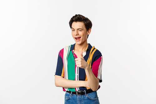Handsome young gay man showing thumb-up and winking, encourage you, praising nice choice, standing over white background.