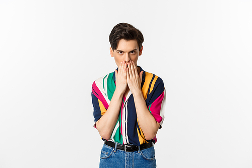 Image of young gay man looking intrigued, holding hands on mouth, gossiping, standing over white background.