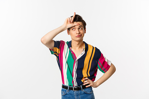 Image of arrogant young man showing loser sign on forehead to mock, looking away with pleased grin, standing over white background.