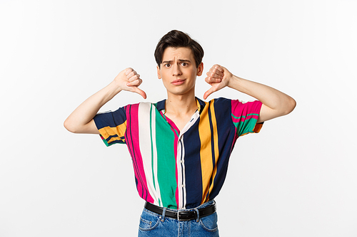 Disappointed queer gay judging something bad, showing thumbs down and grimacing displeased, dislike and disagree, standing over white background.