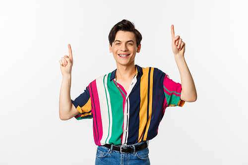 Handsome queer man in stylish clothes showing logo, pointing fingers up and smiling pleased, standing over white background.