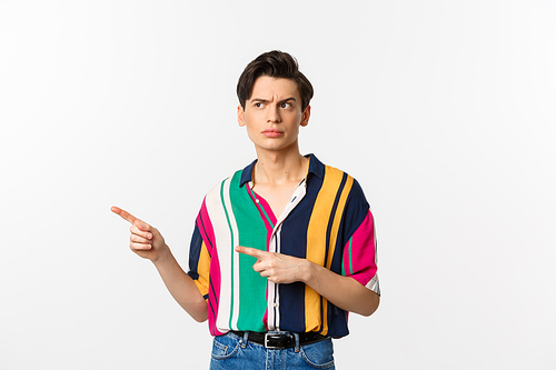 Suspicious young man looking and pointing fingers left with doubtful face, standing unsure in summer shirt, white background.