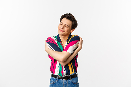 Happy young man hugging himself with eyes closed, smiling pleased, dreaming about something, standing over white background.