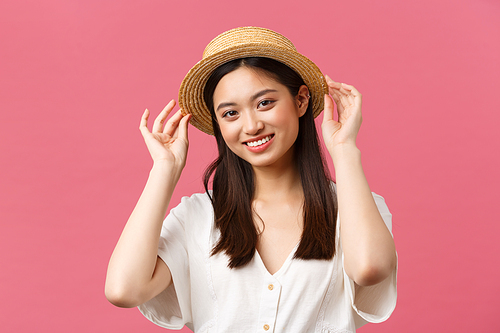 Beauty, people emotions and leisure and vacation concept. Lovely asian woman shopping in store, picking new straw hat, smiling delighted, buying summer outfit over pink background.