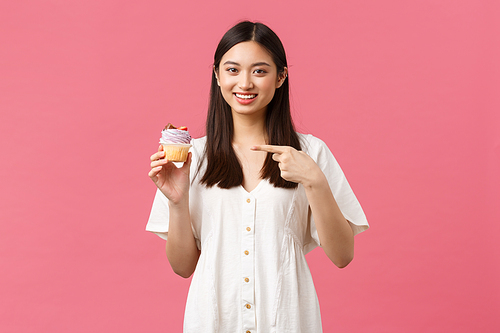 Food, cafe and restaurants, summer lifestyle concept. Happy asian woman satisfied with awesome taste of dessert, pointing at cupcake recommend bakery shop, standing pink background.