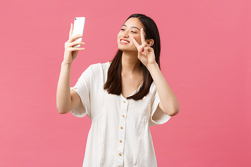 Beauty, people emotions and technology concept. Feminine good-looking stylish asian girl blogger taking selfie on smartphone camera, smiling happy at mobile phone, standing pink background.