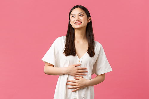 Beauty, people emotions and summer leisure and vacation concept. Pleased smiling asian girl looking dreamy as sky and rubbing belly, got rid of cramps or stomach ache with medicine, pink background.