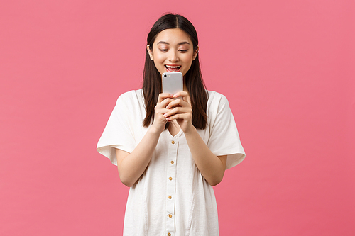 Beauty, emotions and technology concept. Excited asian girl blogger looking at mobile phone with astonished happy face, texting or using application, watching video on smartphone, pink background.