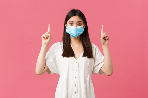 Covid-19, social distancing, virus and lifestyle concept. Pretty stylish asian girl in medical mask suggest visit this store or place, pointing fingers up at product, standing pink background.