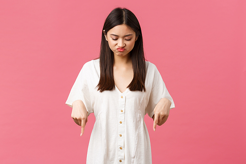 Beauty, people emotions and summer leisure and vacation concept. Disappointed and upset cute gloomy asian girl pointing and looking down with regret and sadness, pink background.