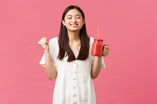 Healthy food, emotions and summer lifestyle concept. Enthusiastic and cheerful, smiling asian woman boosting energy with delicious fresh smoothie, drinking and looking upbeat, pink background.