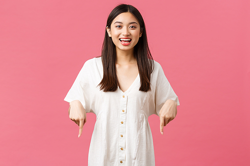 Beauty, people emotions and summer leisure and vacation concept. Joyful pretty korean girl in white dress pointing fingers down and smiling excited, demonstrate awesome new promo offer.