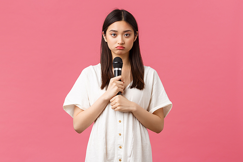 Leisure, people emotions and lifestyle concept. Gloomy and reluctant young asian girl holding microphone and looking sad camera, unwilling to perform, standing moody pink background.