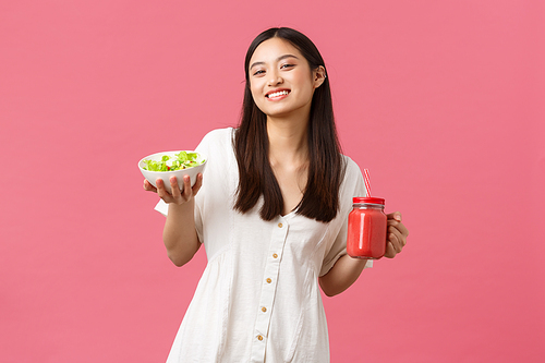 Healthy food, emotions and summer lifestyle concept. Enthusiastic and upbeat cute asian girl full of energy, eating tasty fresh salad and drinking smoothie, smiling at camera happy, pink background.