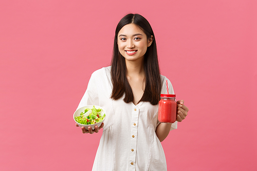 Healthy food, emotions and summer lifestyle concept. Good-looking young 20s woman taking care of body, eating salad and drinking fresh smoothie, standing pink background.